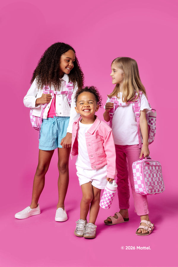 Three children wearing coordinated pink, white, and blue outfits stand against a hot pink background, holding and wearing Simple Modern branded backpacks and accessories with pink and white checkered patterns.