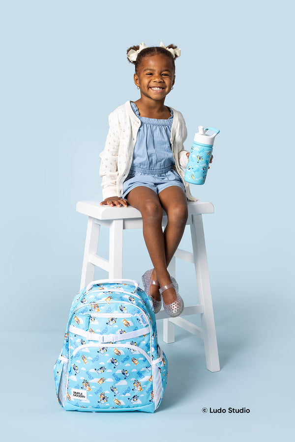 Young girl sitting on a white stool wearing a light blue denim dress and cream cardigan, holding a cyan Bluey water bottle with a matching blue Bluey backpack at her feet, against a soft blue background.