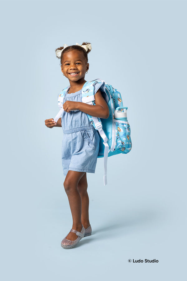 Young girl wearing light blue chambray dress with white hair bows, sparkly lavender Mary Jane shoes, and turquoise Bluey backpack with white accents, smiling at camera against light blue background.