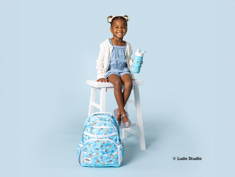 Young child with dark skin smiling at camera, sitting on white stool wearing light blue denim romper and cream cardigan. Child holds light blue water bottle and matching backpack with colorful character illustrations against