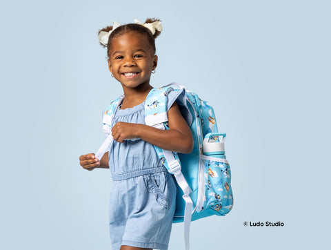 Child wearing pale blue outfit with cream hair bows smiling while carrying a turquoise Bluey character backpack against a light blue studio background.