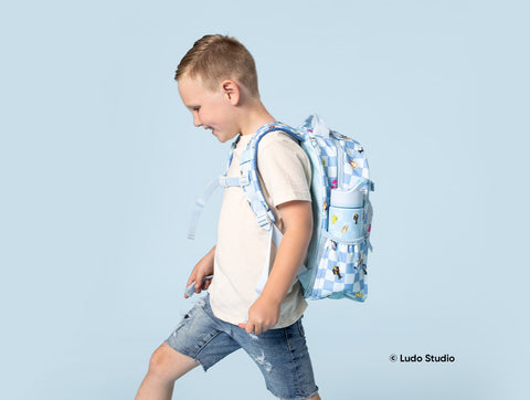 Young boy in profile wearing a light blue Bluey-themed backpack with checkered pattern and character prints, paired with a cream t-shirt and denim shorts, smiling against a pale blue studio background.