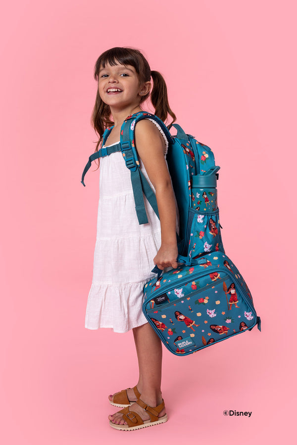 Young girl smiling at camera wearing white tiered dress, holding teal Disney Moana lunch bag, and wearing matching Moana backpack against pink background.
