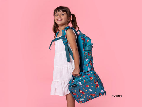Young girl in white tiered dress smiling over shoulder, wearing teal blue Disney backpack and holding matching lunch bag with holiday character prints on pink background.