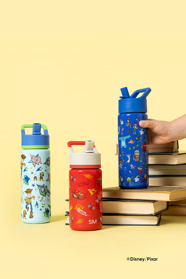 Three Simple Modern kids water bottles featuring Disney-Pixar characters: mint green Toy Story bottle on left, bright red Cars bottle in center, and royal blue Toy Story bottle on right, displayed on stacked books against