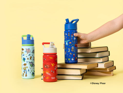 Three Simple Modern water bottles featuring Disney and Pixar characters in blue, red, and mint green colors arranged in front of stacked books on a pale yellow background, with a hand reaching for the blue bottle.