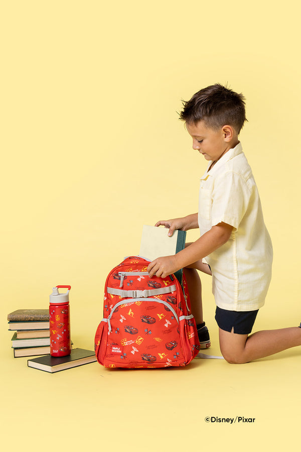 Young boy kneeling beside a red Disney-Pixar backpack on pale yellow background, with stacked books and red Simple Modern water bottle, showcasing school supplies in studio setting.