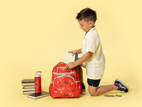 Young boy kneeling and reaching toward a red Disney-Pixar backpack with colorful icons, surrounded by stacked textbooks and a red water bottle on a pale yellow background.