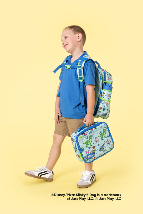 Young boy wearing blue t-shirt and khaki shorts holding a blue Simple Modern backpack and matching lunch bag decorated with Disney-Pixar cartoon characters against a pale yellow background.