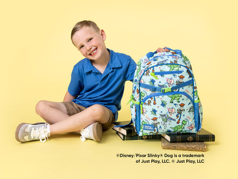 Child in blue shirt sitting cross-legged with Disney-Pixar Toy Story backpack on yellow studio background, featuring Woody and Buzz Lightyear characters on cream and blue pack with decorative books beneath.