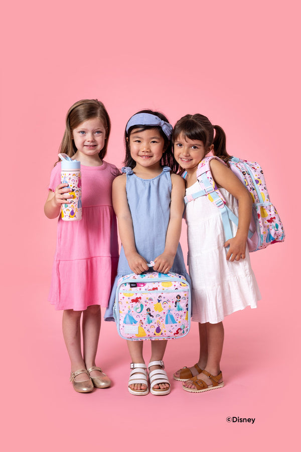 Three smiling girls in pastel clothing holding Disney princess-themed Simple Modern products - a water bottle, lunch box, and backpack - against a bright pink background.