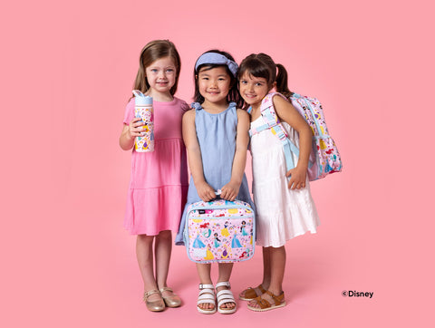 Three smiling girls holding Disney Princess-themed Simple Modern products including a water bottle, lunch box, and backpack against a pink background.
