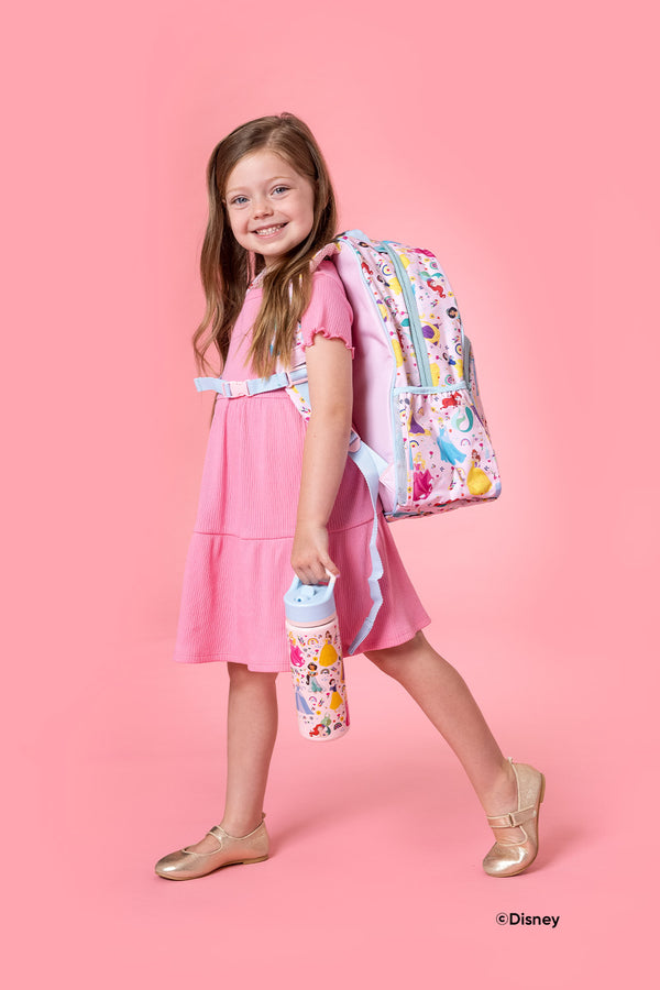 Young girl wearing a light pink ribbed dress with blue belt, gold ballet flats, holding a Disney Princess-themed backpack and water bottle against a soft pink background.