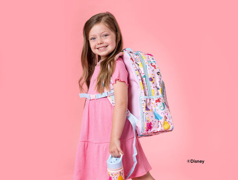 Young girl wearing pink outfit smiling at camera, holding light blue water bottle and wearing matching backpack, both featuring Disney Princess characters on soft pink studio background.