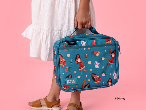 Child holding teal Simple Modern lunch box featuring Disney Princess characters, flowers, and stars against pink background. Child wears white tiered dress and tan sandals.