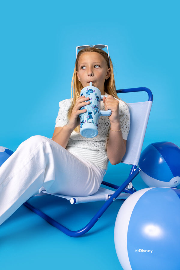 Young girl with reddish-blonde hair sitting on a blue lounge chair, drinking from a Simple Modern water bottle featuring Disney Stitch graphics. She wears white clothing and sunglasses on her head against a bright turquoise