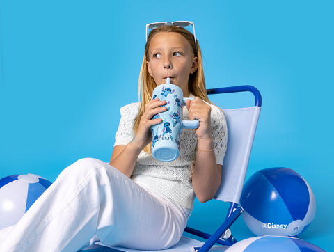 Young girl seated on blue and white chair holding Simple Modern water bottle with Stitch graphics, wearing white sunglasses, gray cardigan, and white pants, surrounded by beach balls on bright blue background.