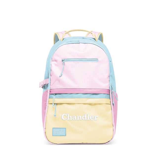 Simple Modern Chandler colorblock backpack in pastel pink, blue, and yellow horizontal stripes with front zippered pocket, padded shoulder straps, and embroidered branding on white background.