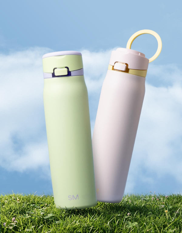 Two pastel water bottles on green grass with blue sky background: left bottle in pale yellow-green with lavender band, right in blush pink with gold accents. Easter-themed outdoor lifestyle product image.