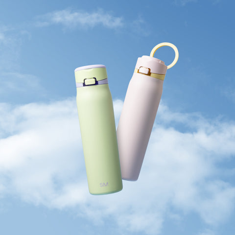 Two Simple Modern water bottles in soft green and light pink with white caps, displayed against a dreamy blue sky with white clouds. Easter spring collection product photography.