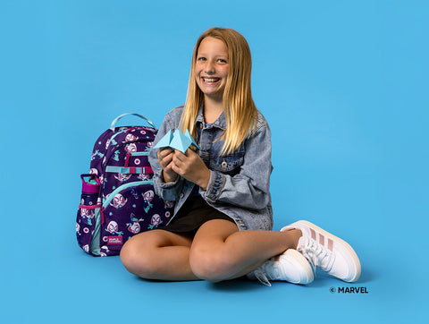 Young girl with strawberry-blonde hair smiling at camera, wearing light blue denim jacket and white sneakers, holding a light blue lunch box. Marvel-licensed backpack with purple base and colorful character designs sits