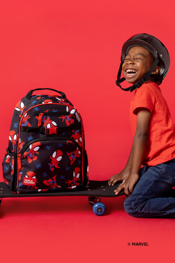 Young boy wearing black helmet and orange-red shirt, kneeling happily against coral-red background with Spider-Man backpack and blue skateboard wheel visible.