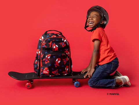 Child sitting on black skateboard wearing protective helmet and orange shirt, smiling at camera. Marvel-themed Simple Modern backpack with Spider-Man graphics sits beside child against coral-red background.
