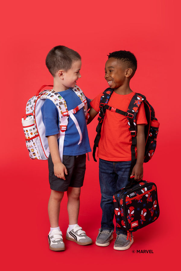 Two smiling children facing each other against a bright red background, wearing Marvel-themed backpacks and casual clothing. Product photography showcasing youth-sized school bags and accessories.