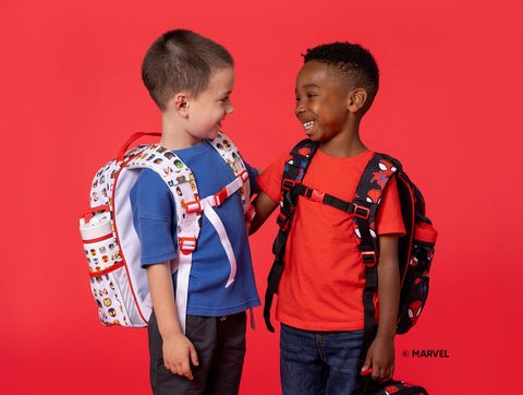 Two smiling elementary school boys wearing Simple Modern backpacks against a bright red background. Left boy in blue shirt with food-themed backpack, right boy in red shirt with Marvel character backpack.