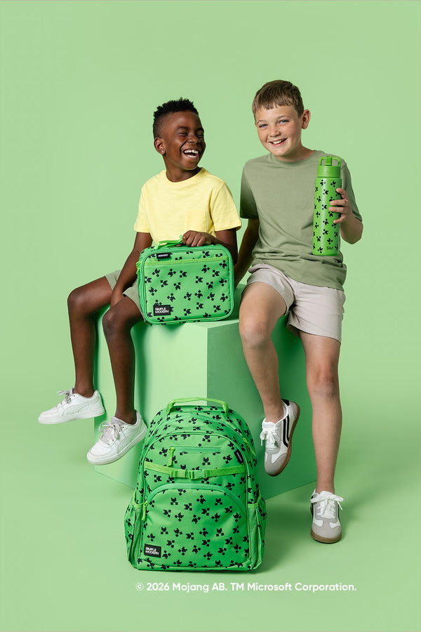 Two children sitting on a lime green platform wearing white sneakers and holding Simple Modern Minecraft collection items: a lunch bag, water bottle, and backpack, all featuring black pixelated designs on lime green fabric.