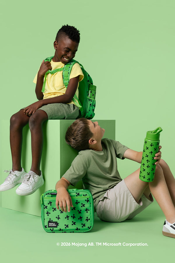 Two smiling children display Simple Modern Minecraft-collection products against a bright green background: one wearing a backpack on an elevated platform, the other holding a lunch box and water bottle.