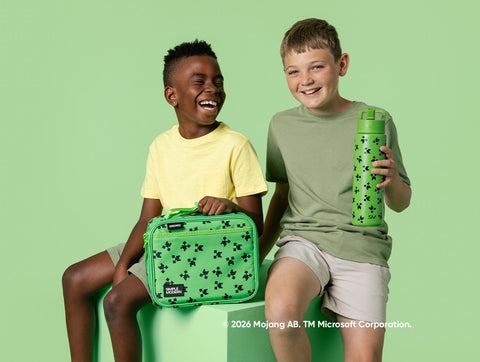 Two smiling children on a green platform holding a matching lime green Minecraft lunch box and water bottle with black cross patterns against a bright green background.