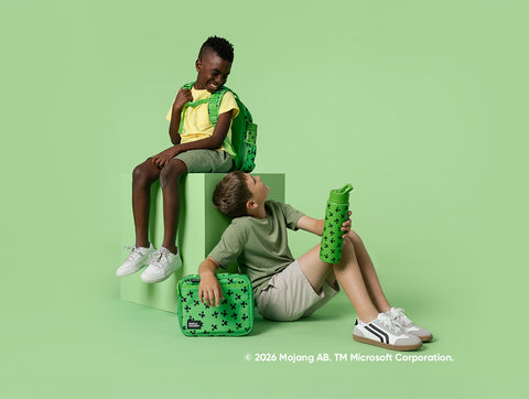 Two children wearing Simple Modern backpacks and holding water bottles on a sage green background. One child sits on a pale green block wearing a yellow shirt, the other sits below wearing a sage green shirt. Minecraft