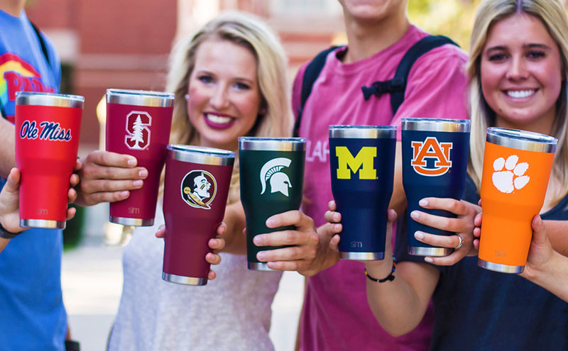 Group of smiling people holding colorful insulated tumblers in red, maroon, navy blue, black, and orange in a bright lifestyle product photo setting.