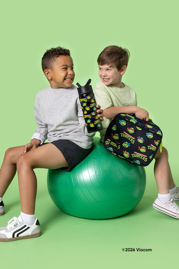 Two boys on a bright green exercise ball holding TMNT-branded products: a black water bottle with turtle graphics and a black lunch bag with 