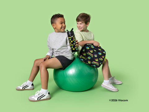 Two smiling boys sitting on a green exercise ball holding TMNT-branded water bottle and lunch bag against a bright lime green background.