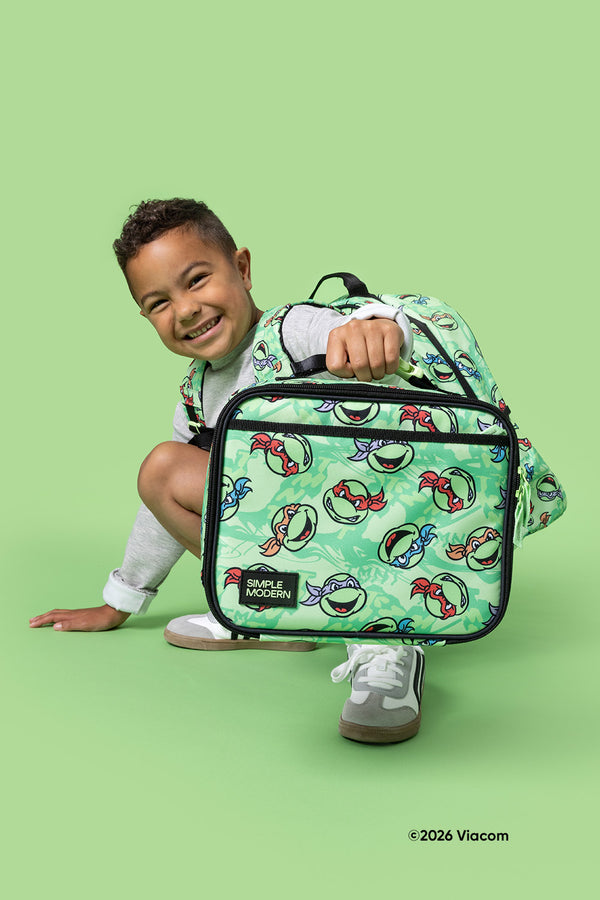 Child in gray shirt crouching and smiling while holding a sage green TMNT-themed insulated lunch bag with matching backpack against a bright mint green background.