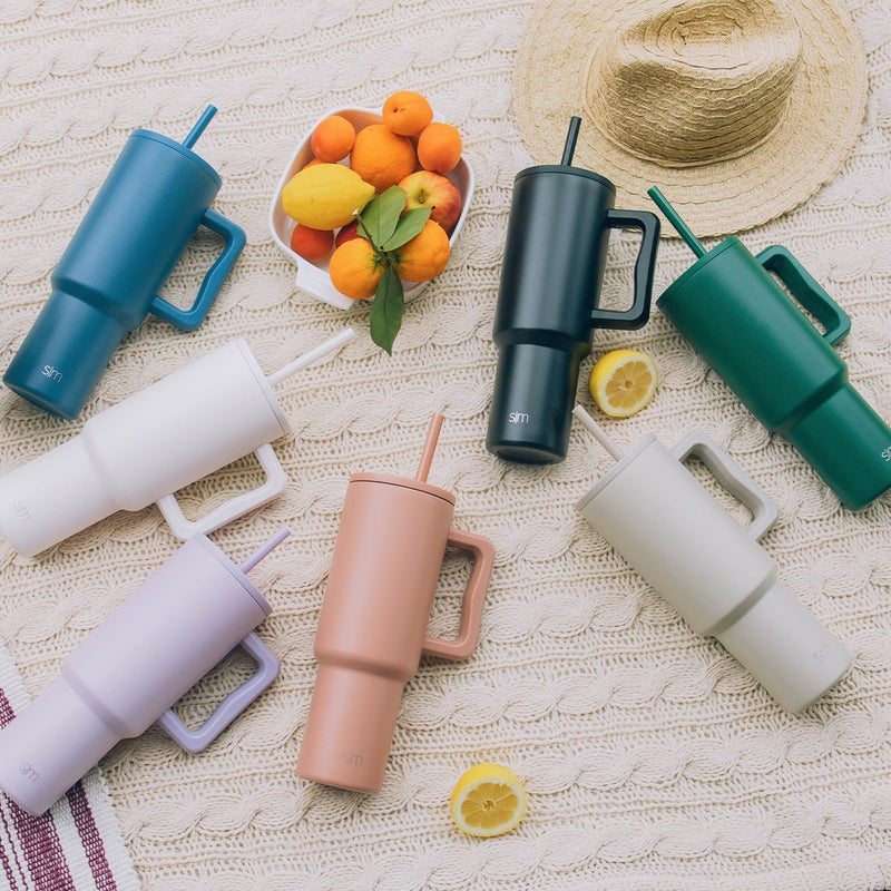 Flat lay of summer essentials including a cream textured fabric, woven straw hat, teal blue sunglasses, forest green and soft pink items, and lavender accents on a white background.