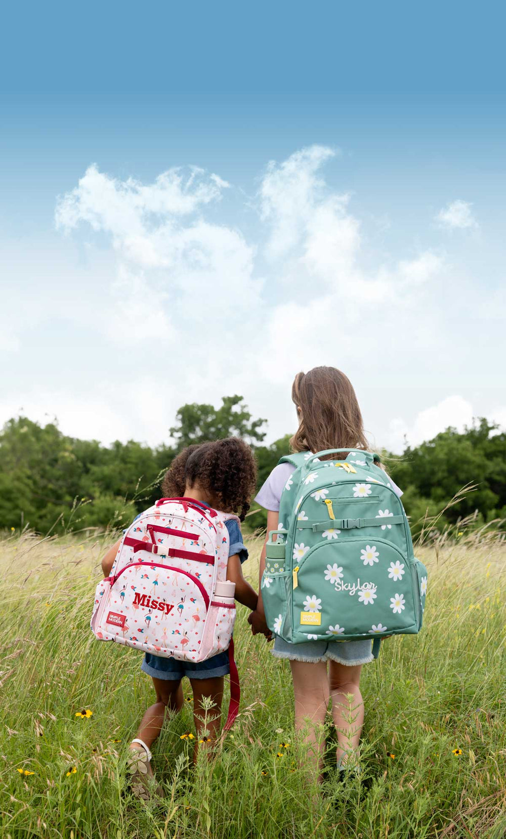 Two children walking through a grassy field wearing personalized backpacks.