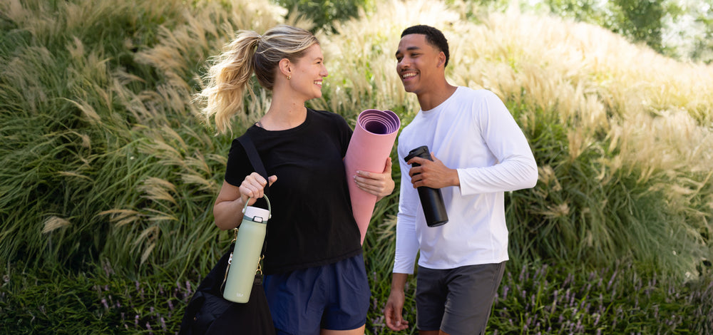 Two friends walking a garden path beside tall ornamental grasses, one carrying a yoga mat and a mint Simple Modern bottle, the other a black bottle.