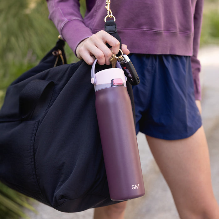 A plum bottle carried by its handle alongside a black duffel bag on a garden path.