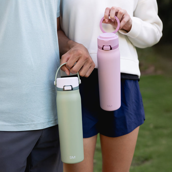Two people standing on grass, each carrying a bottle by its handle - one sage green, one pink.