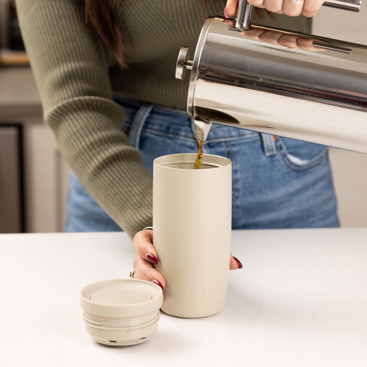 Pouring coffee from a French press into a cream travel tumbler on a kitchen counter